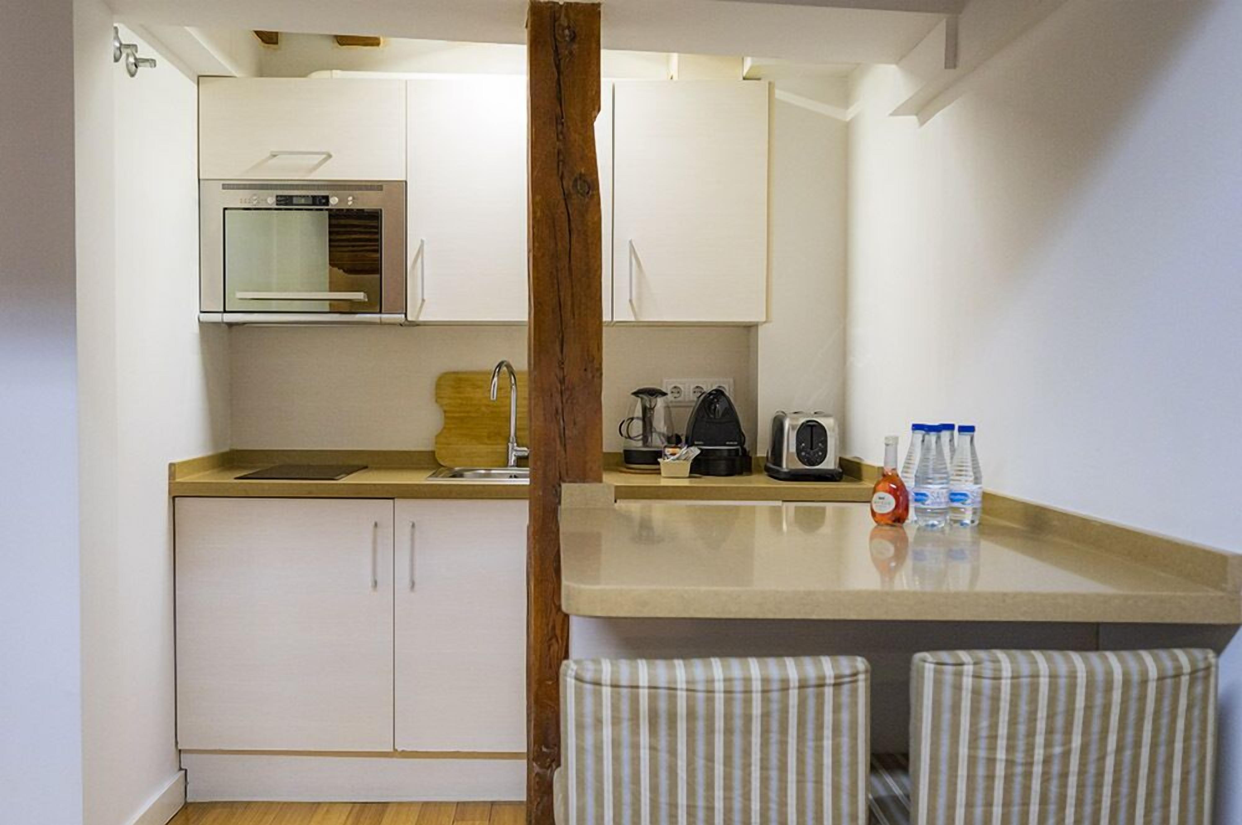 Een compacte keuken met een inbouwoven, een koffiezetapparaat en een waterkoker op het aanrecht. Twee gestreepte stoelen en een houten paal staan in het midden.