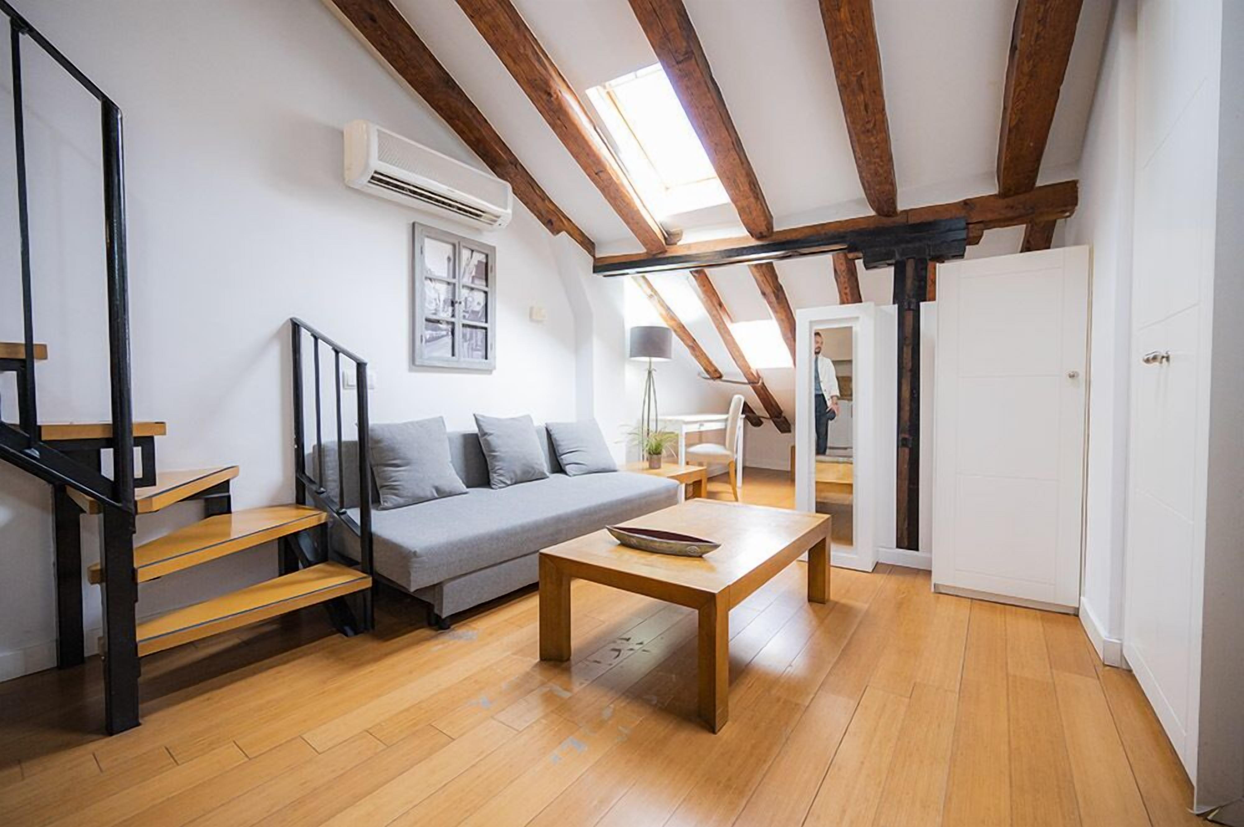 A modern loft with exposed wooden beams, a grey sofa, a wooden coffee table, and a staircase with black railings.