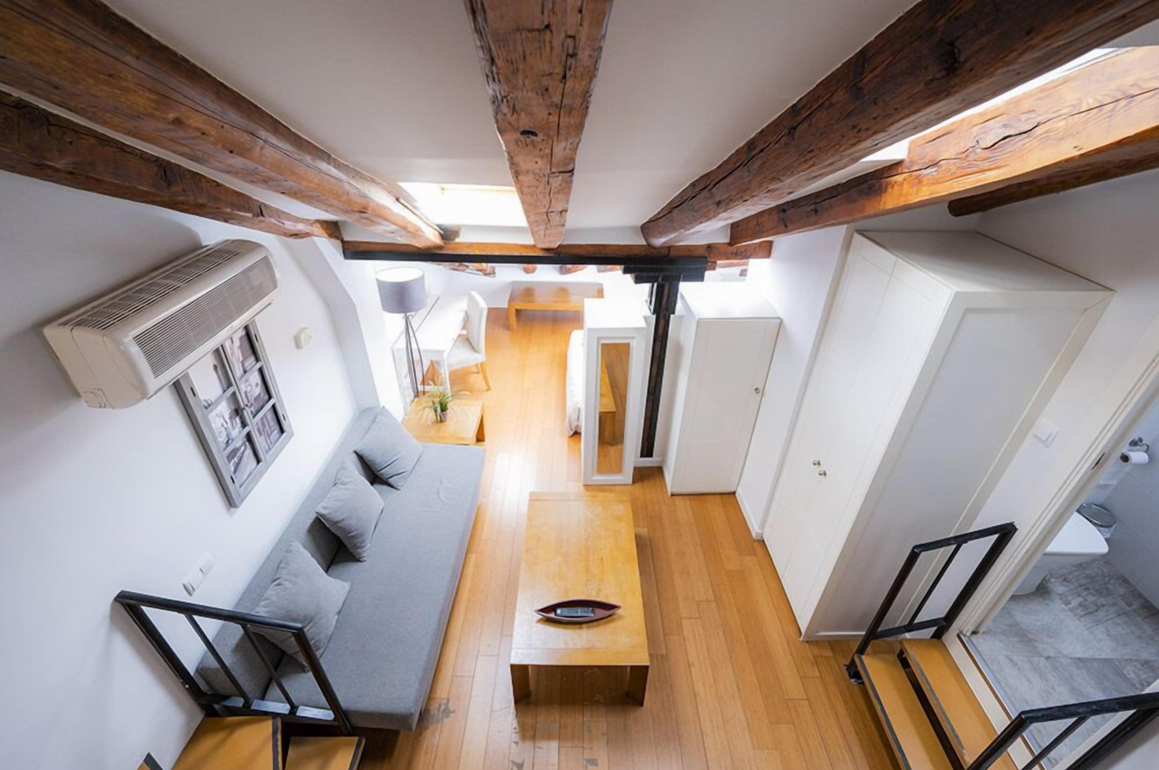 Un intérieur moderne avec des poutres apparentes, une grande table en bois au centre, un canapé gris et un escalier avec des rampes noires.