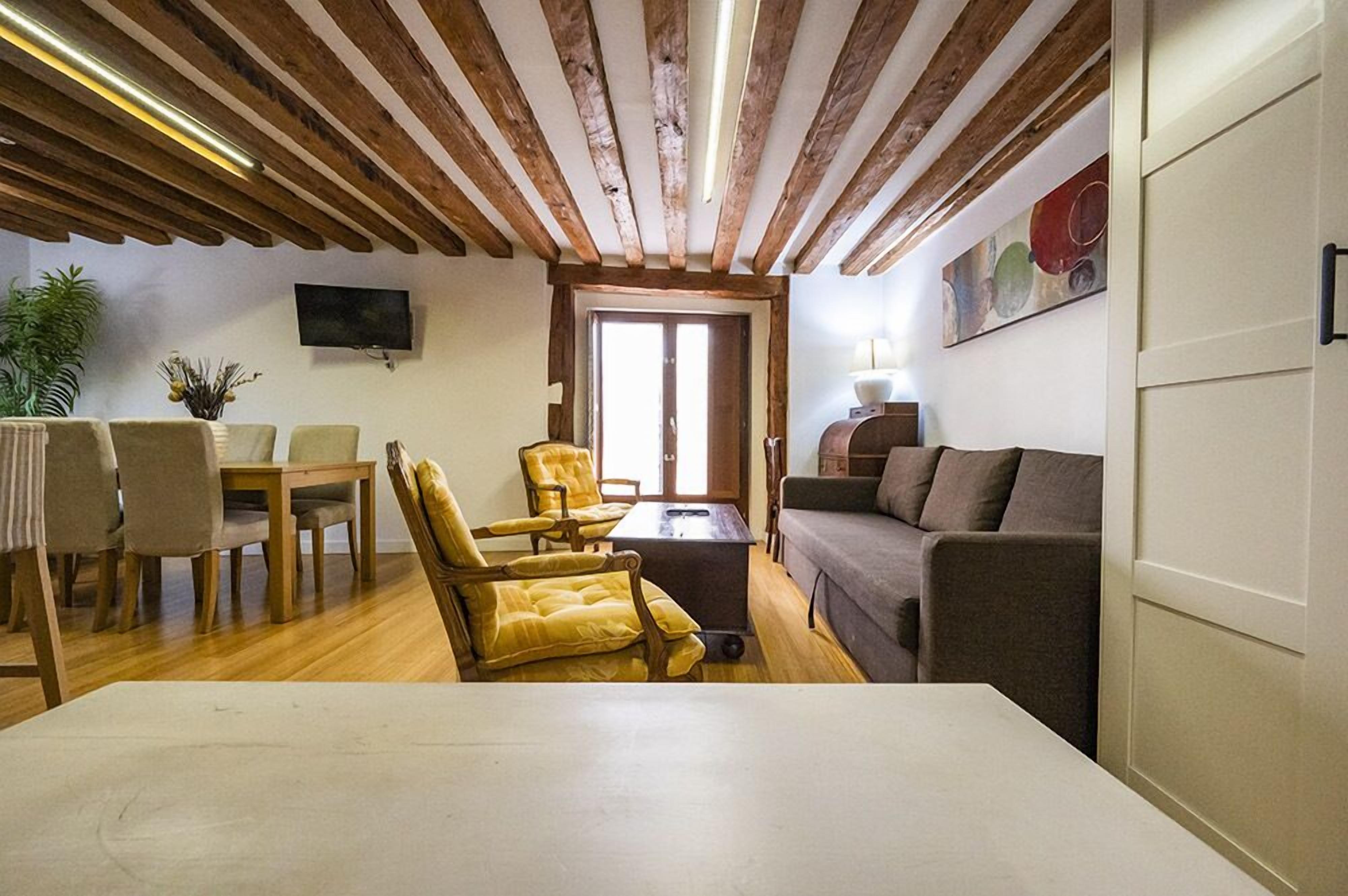 A cozy living room with wooden beams, a sofa, a dining table with chairs, and a TV mounted on the wall.