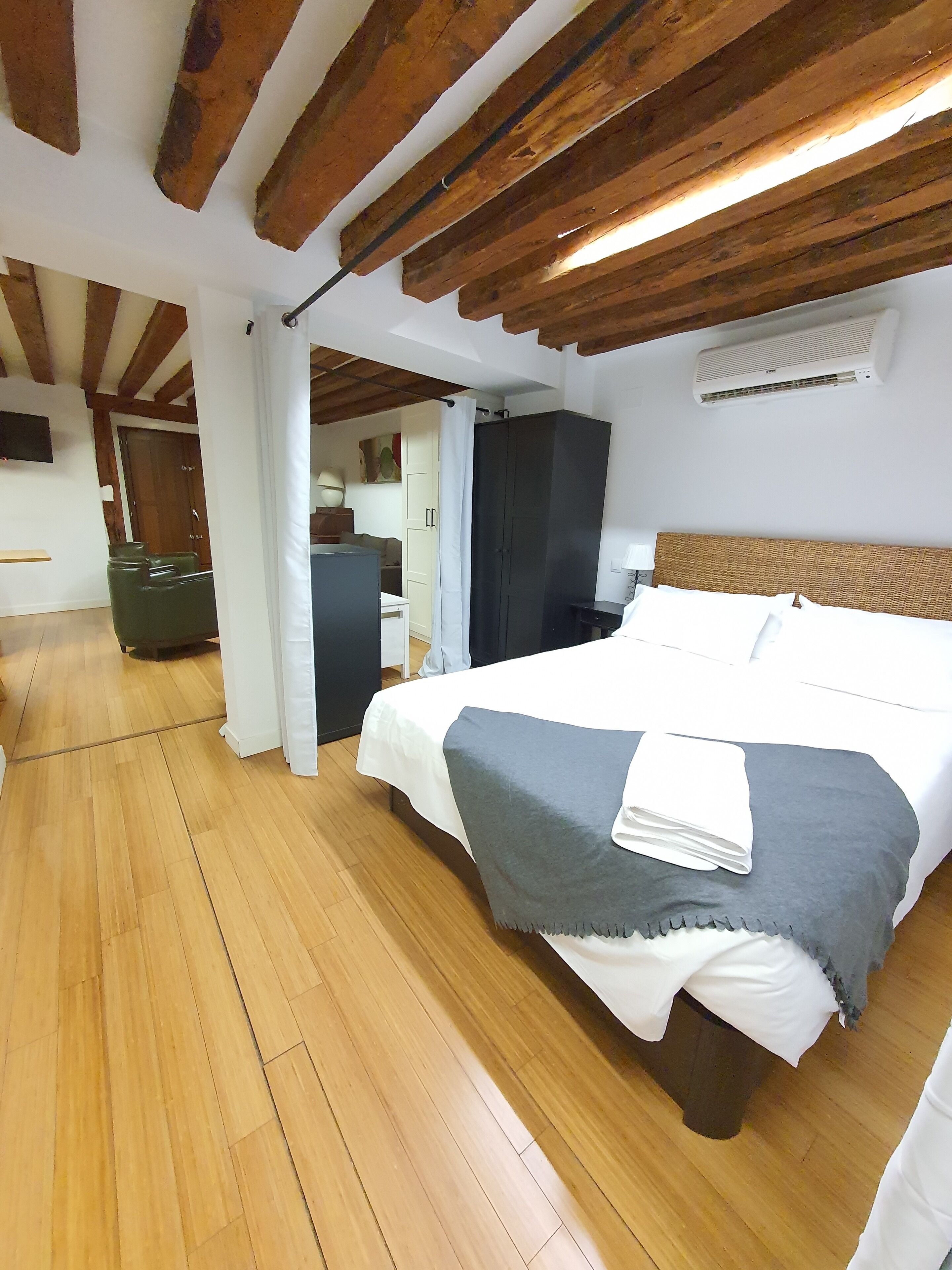 A bedroom with a wooden ceiling, a bed with a grey and white bedspread, a wooden floor, a wardrobe, and a television on the wall.