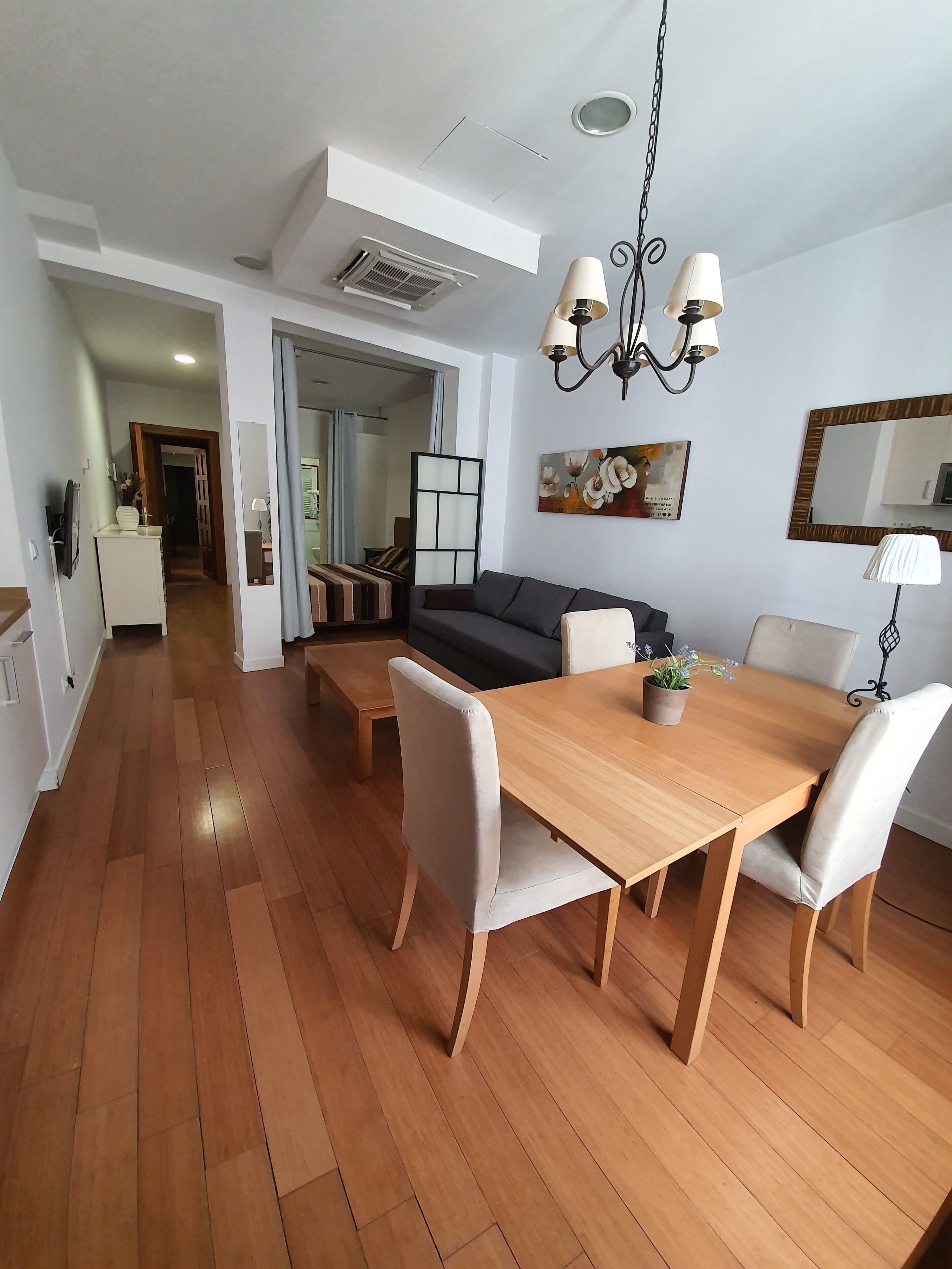 A modern living room with a wooden dining table, white upholstered chairs, a dark sofa, and a chandelier.