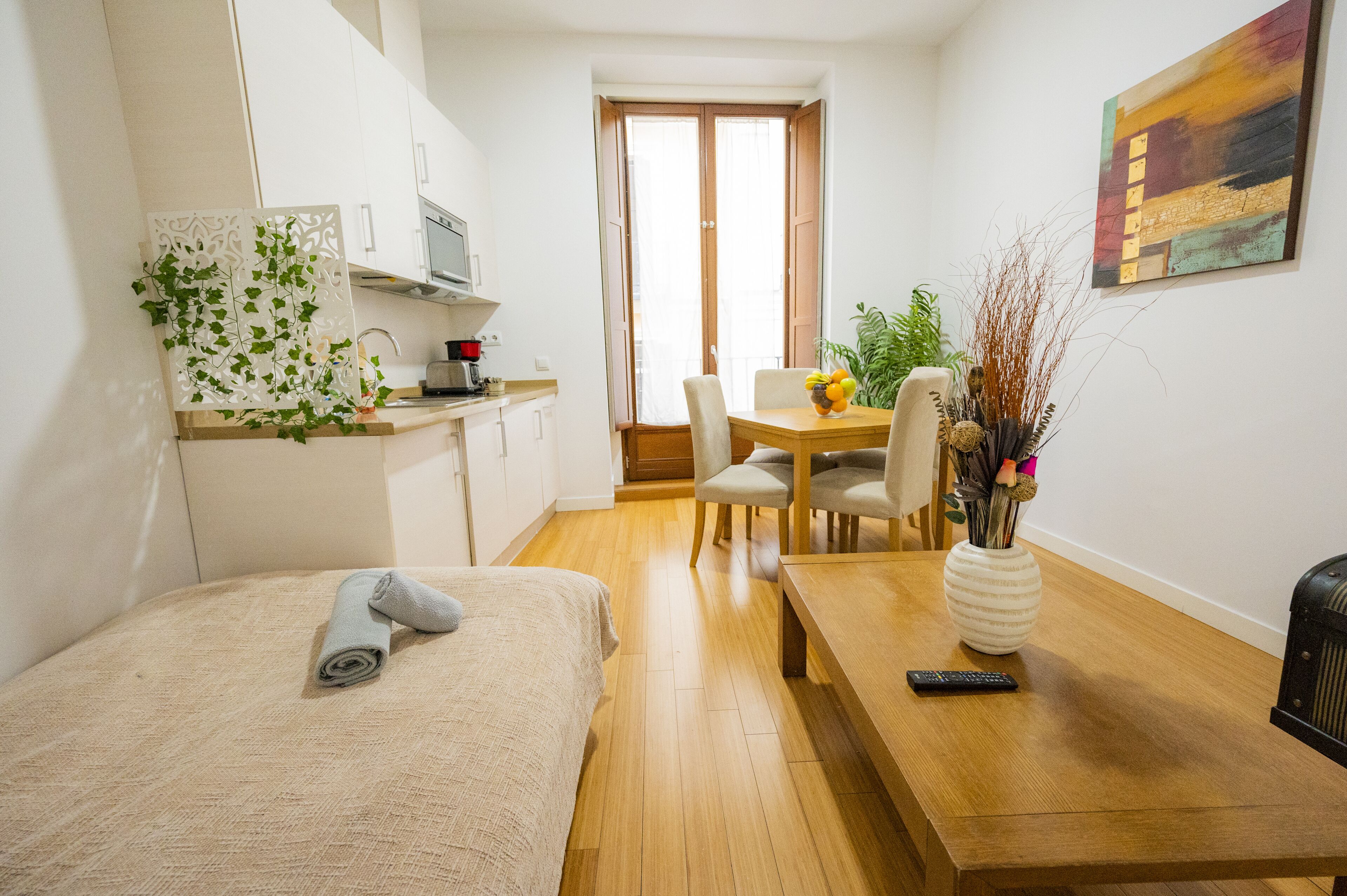 A compact hotel room with a bed, a small kitchenette, a dining area with a table and chairs, and a painting on the wall.