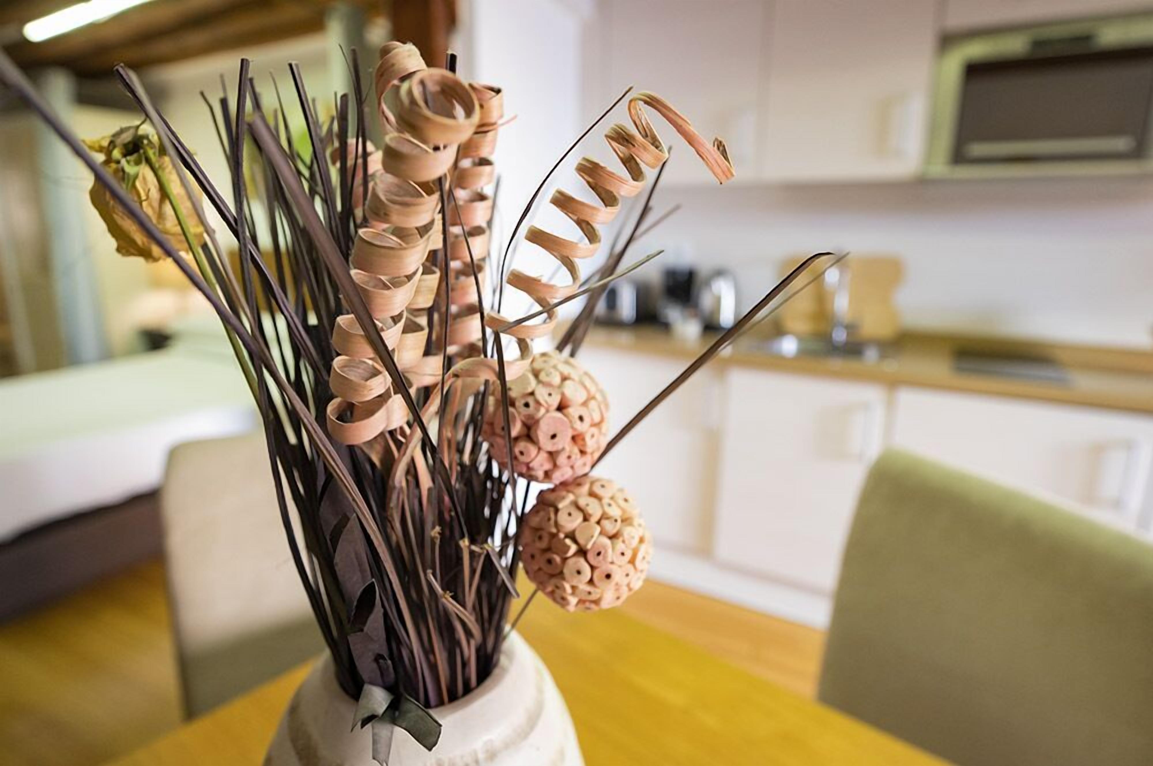 Un vase contenant des plantes séchées et des éléments décoratifs, posé sur une table dans une pièce où l’on aperçoit une cuisine en arrière-plan.