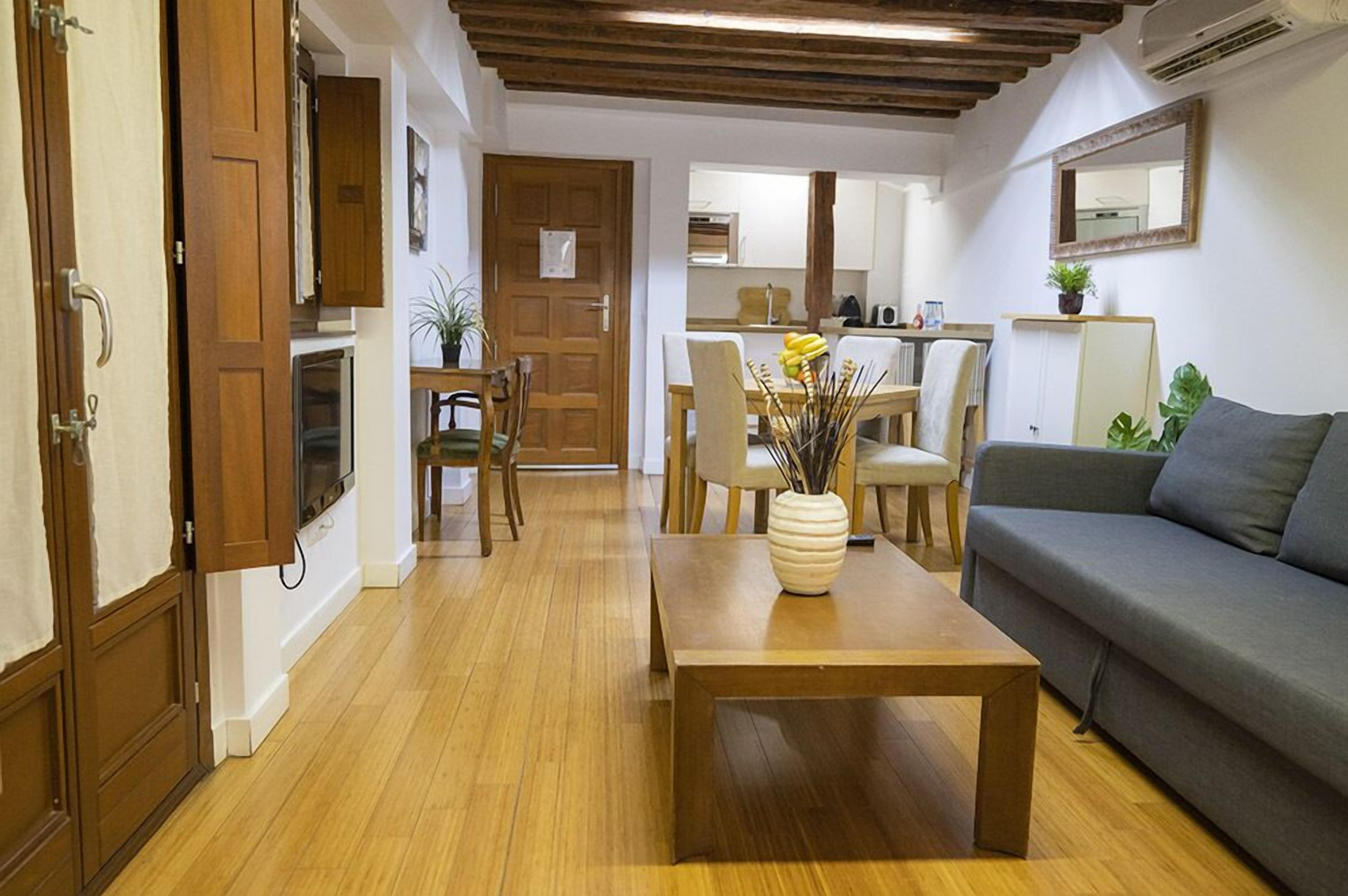 A cozy living room with a wooden floor, a grey sofa, a wooden coffee table with a vase, and a dining area with chairs and a table.