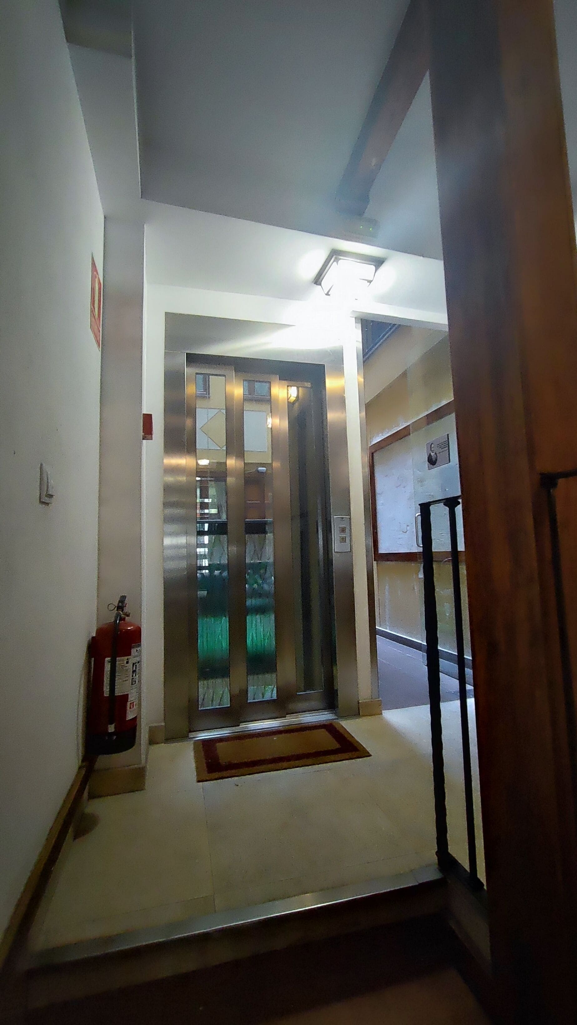 A hallway with an elevator, a fire extinguisher, and a door with a security camera.