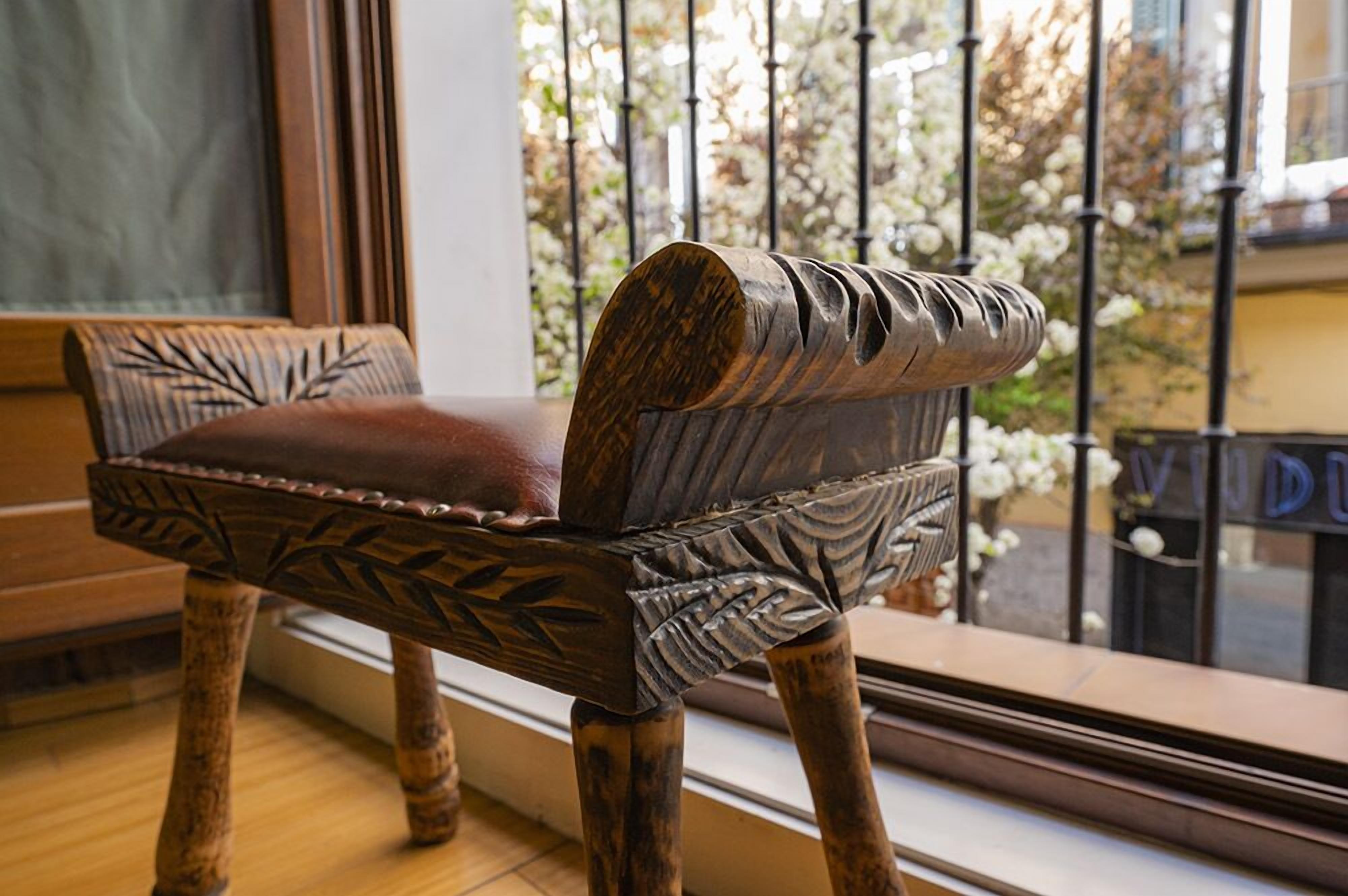 Un banc en bois orné de motifs sculptés, placé près d’une fenêtre donnant sur un balcon et des fleurs blanches.