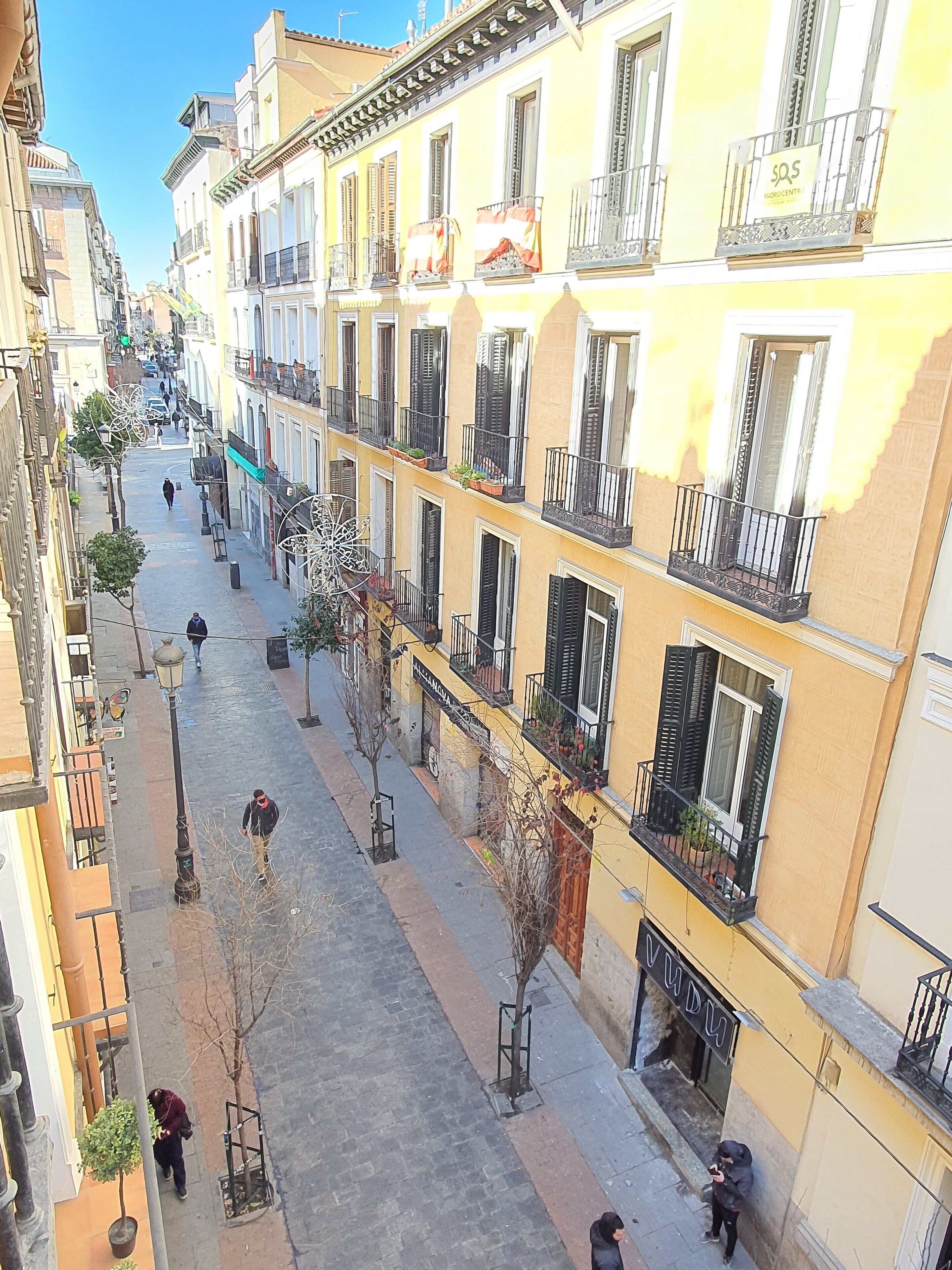 Una calle estrecha flanqueada por edificios de varios pisos, con balcones adornados con plantas y mesas al aire libre.