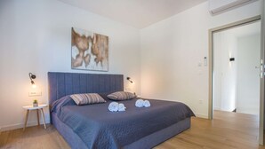 Une chambre moderne avec un grand lit, deux lampes de chevet, une petite table ronde ornée d’une plante et un tableau accroché au mur.