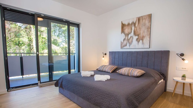 Une chambre moderne avec un grand lit, une porte-fenêtre coulissante donnant sur un balcon et un tableau accroché au mur.