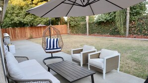 Un patio con una silla mecedora, sofás blancos y una mesa bajo una sombrilla.