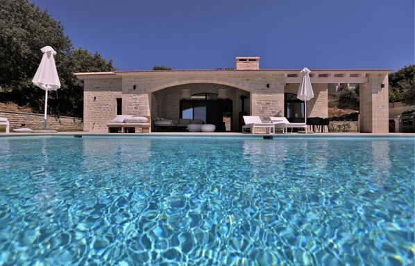 A swimming pool with lounge chairs and umbrellas, a stone building with an arched entrance, and a clear blue sky. - new natural stone villa with large pool and great sea views (Prines)