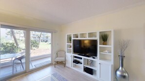 A modern living room with a large flat-screen TV, white shelving, a dining area with a table and chairs, and a view of a building and greenery outside. - Beach and Tennis 118 - Updated Ground floor unit with screen porch and easy (St. Augustine Beach)
