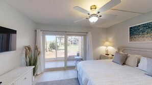 A bedroom with a bed, a ceiling fan, a nightstand with a lamp, a window with curtains, and a view of the outside. - Beach and Tennis 118 - Updated Ground floor unit with screen porch and easy (St. Augustine Beach)
