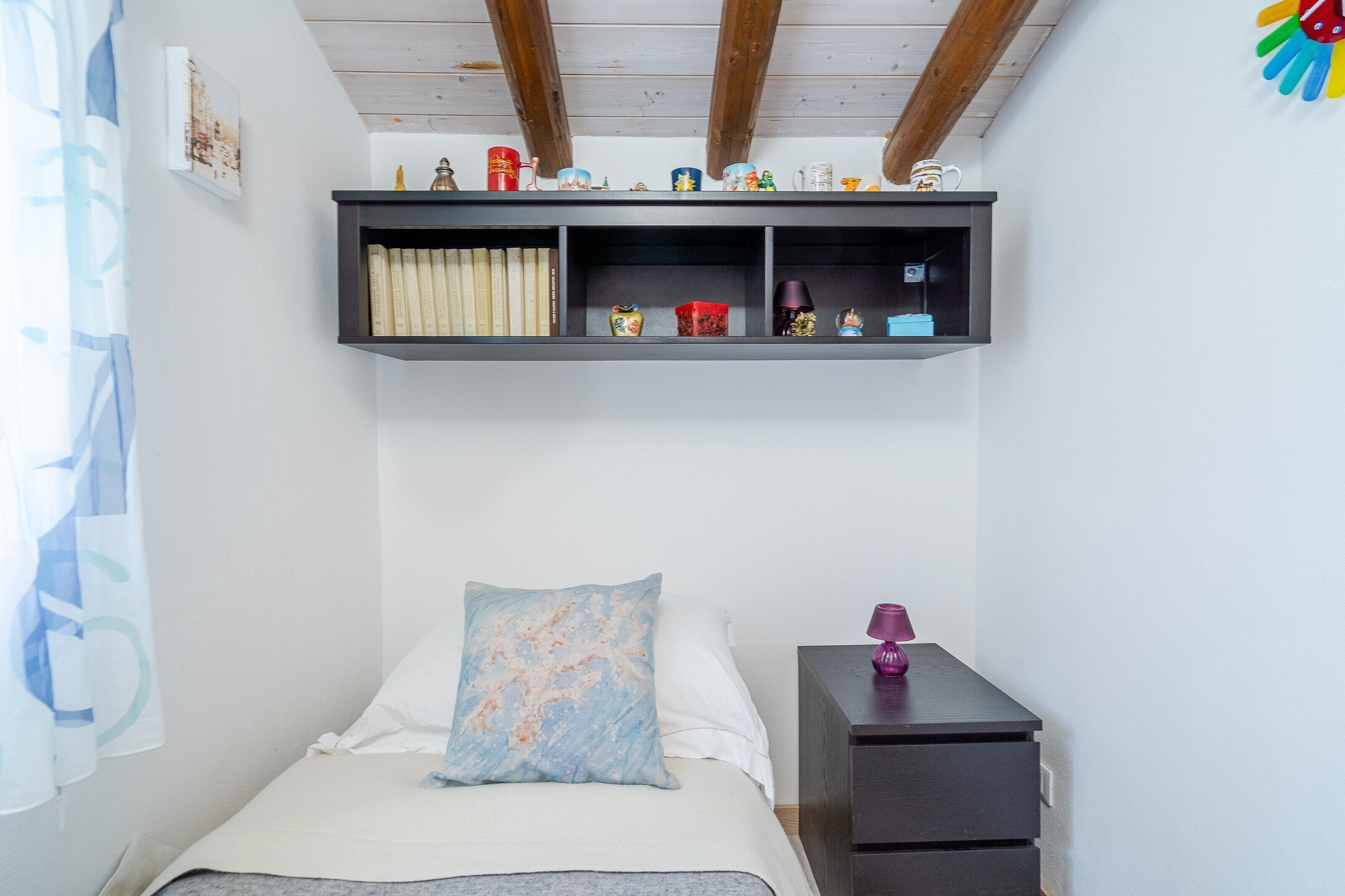 A bedroom with a bed, a nightstand, a shelf with books and decorative items, and a window with curtains.