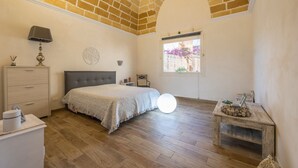 Ein Schlafzimmer mit Bett, Schminktisch, Nachttisch, Lampe, einem Fenster mit Blick und einer Ziegeldecke.