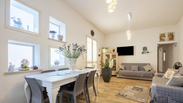 Ein Essbereich mit weißem Tisch und Stühlen, einer großen Kübelpflanze und einer Couch im Hintergrund.