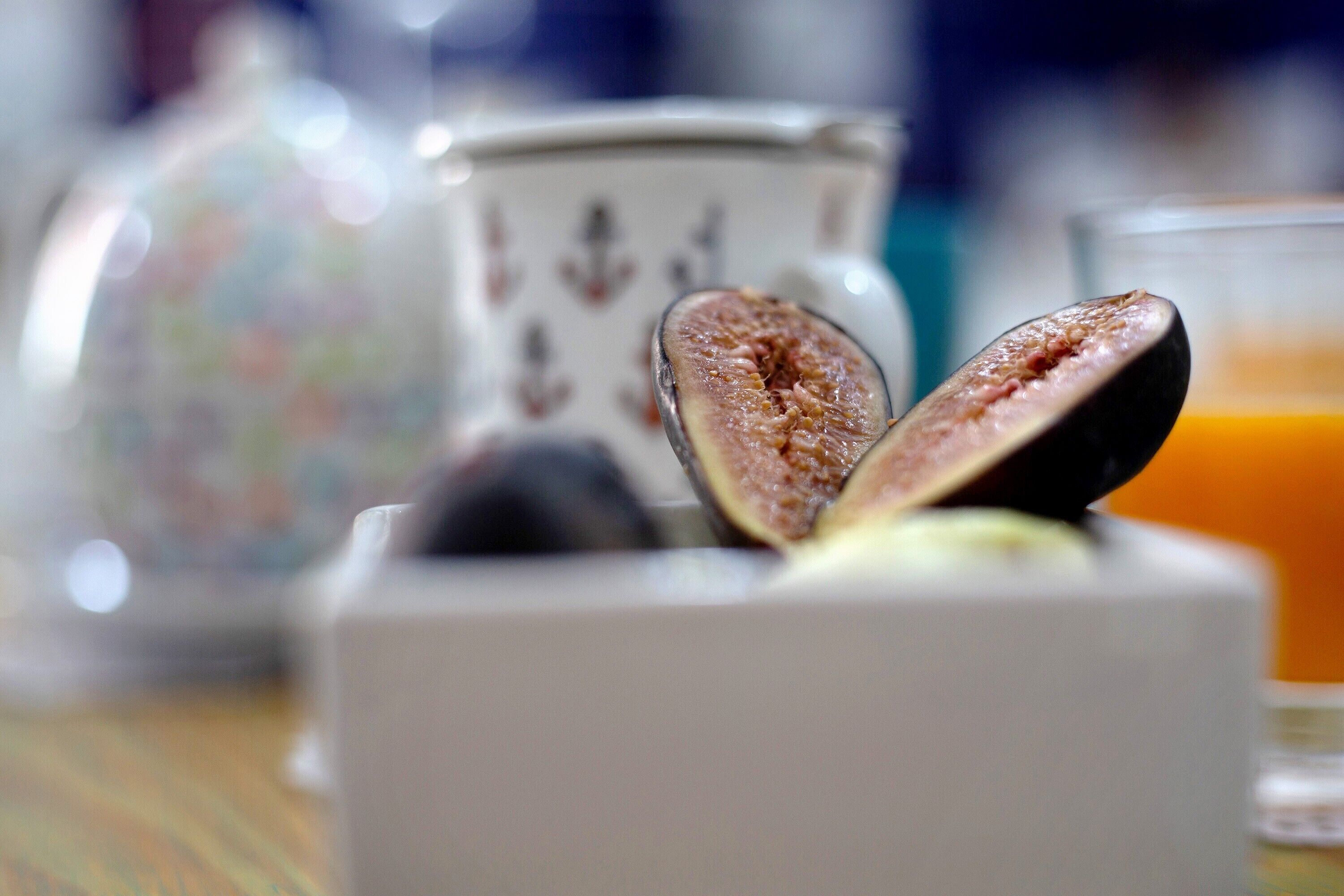 A close-up of a half-cut fig on a white object, with a glass of orange juice and a decorative container in the background.