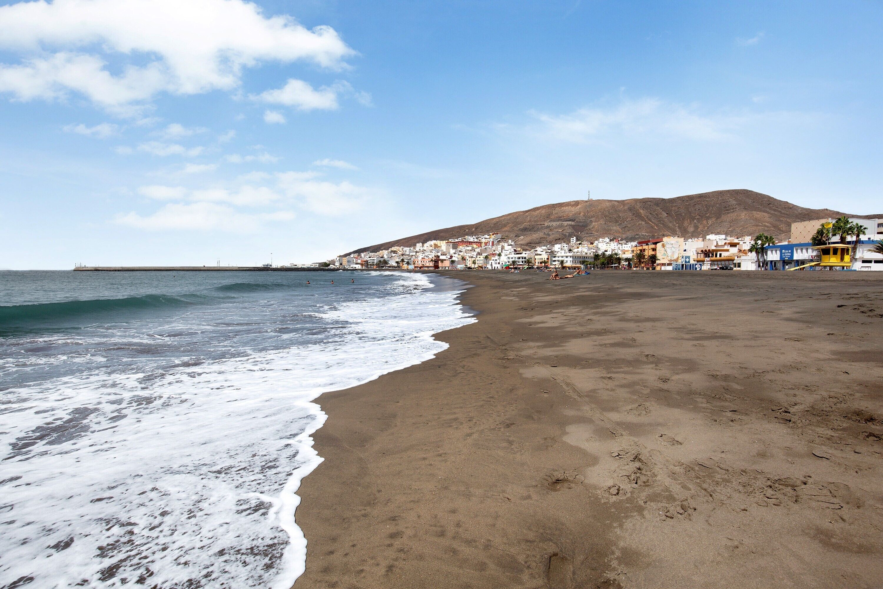 A beach with a mountain in the background, a sandy shore, and buildings along the coastline.