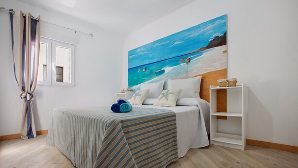 A bedroom with a bed, a painting of a beach scene, a window, and a curtain. - Vacation Home 'Holidays Maxorata Relax' close to the Beach with Wi-Fi (Gran Tarajal)