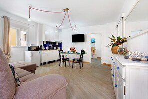 Un salon moderne avec un canapé, un coin repas avec des chaises, une cuisine avec des placards, un téléviseur fixé au mur et une décoration sur le thème de la plage sur la commode.