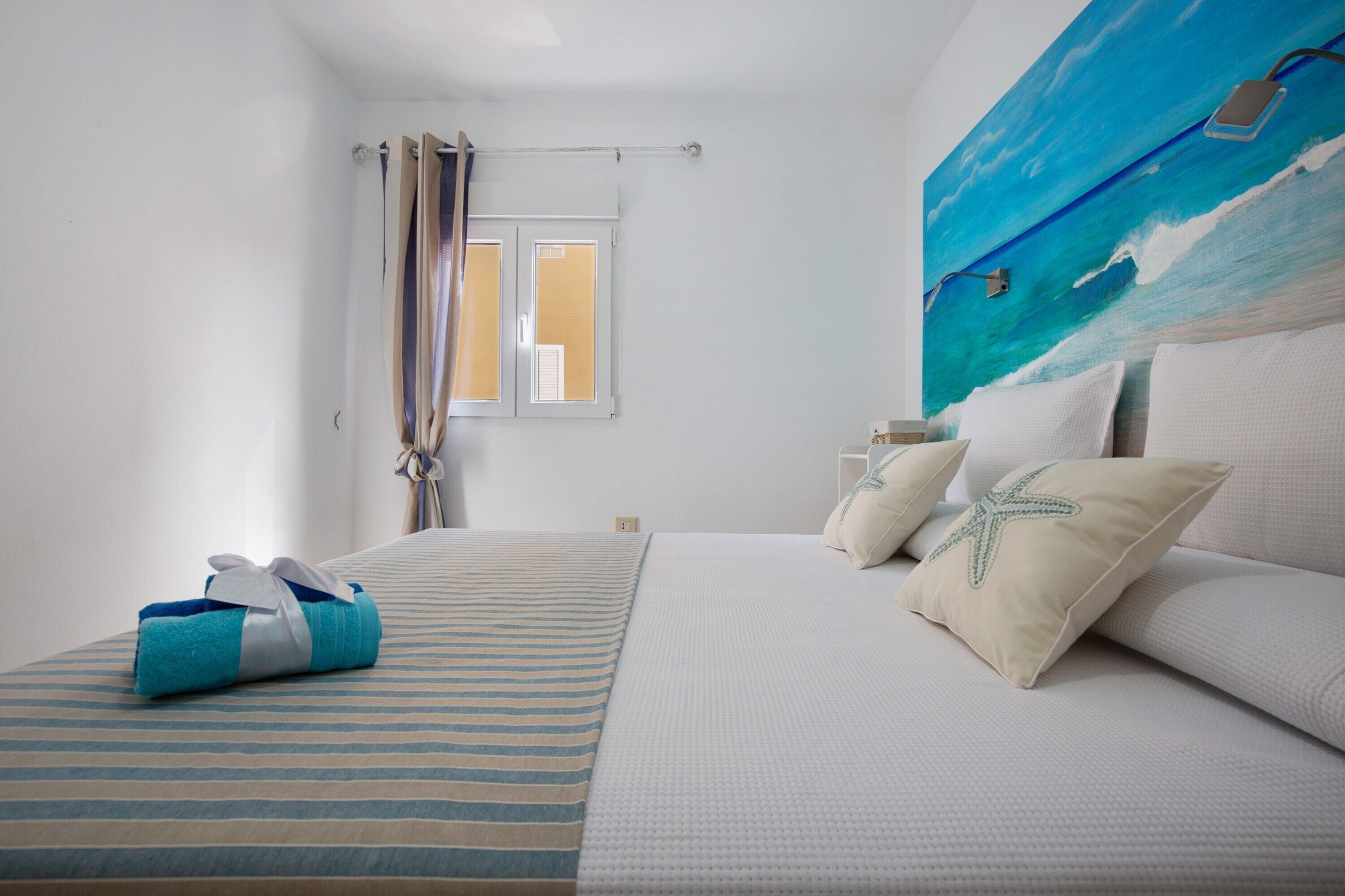 Una camera d'albergo con un letto, una finestra con tende e un quadro che raffigura l'oceano, con un asciugamano sul letto.