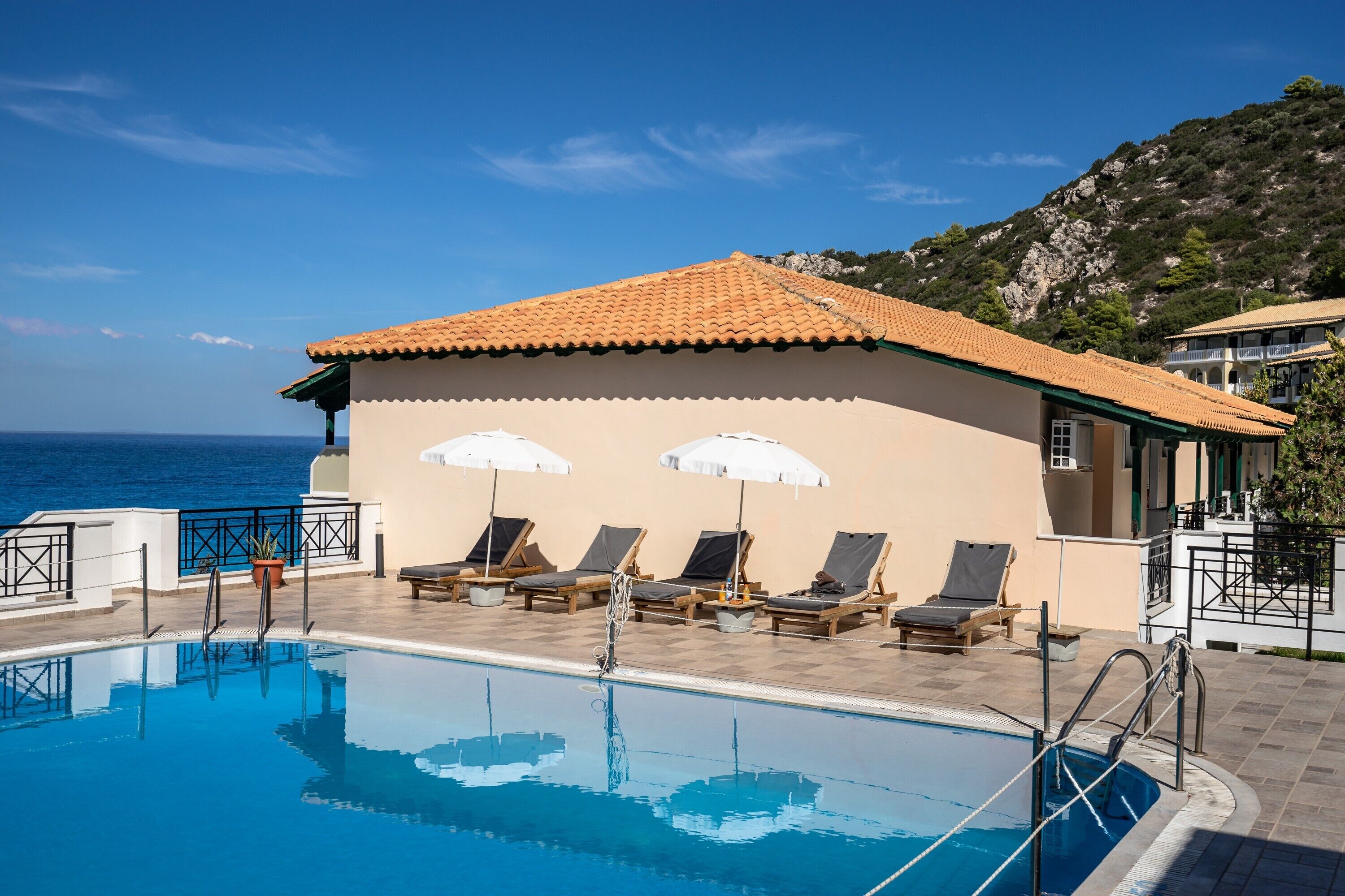 Zona de piscina con sillones de descanso y sombrillas, vista despejada del mar y un edificio con tejado de baldosas.