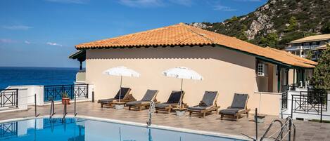 Zona de piscina con sillones de descanso y sombrillas, vista despejada del mar y un edificio con tejado de baldosas.