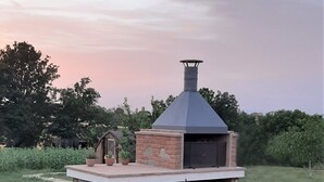 Un barbecue extérieur en briques, doté d’une cheminée, entouré d’une pelouse et de plantes en pot.