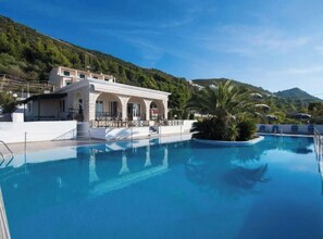 Una piscina con un edificio sullo sfondo, circondata dal verde e da colline.