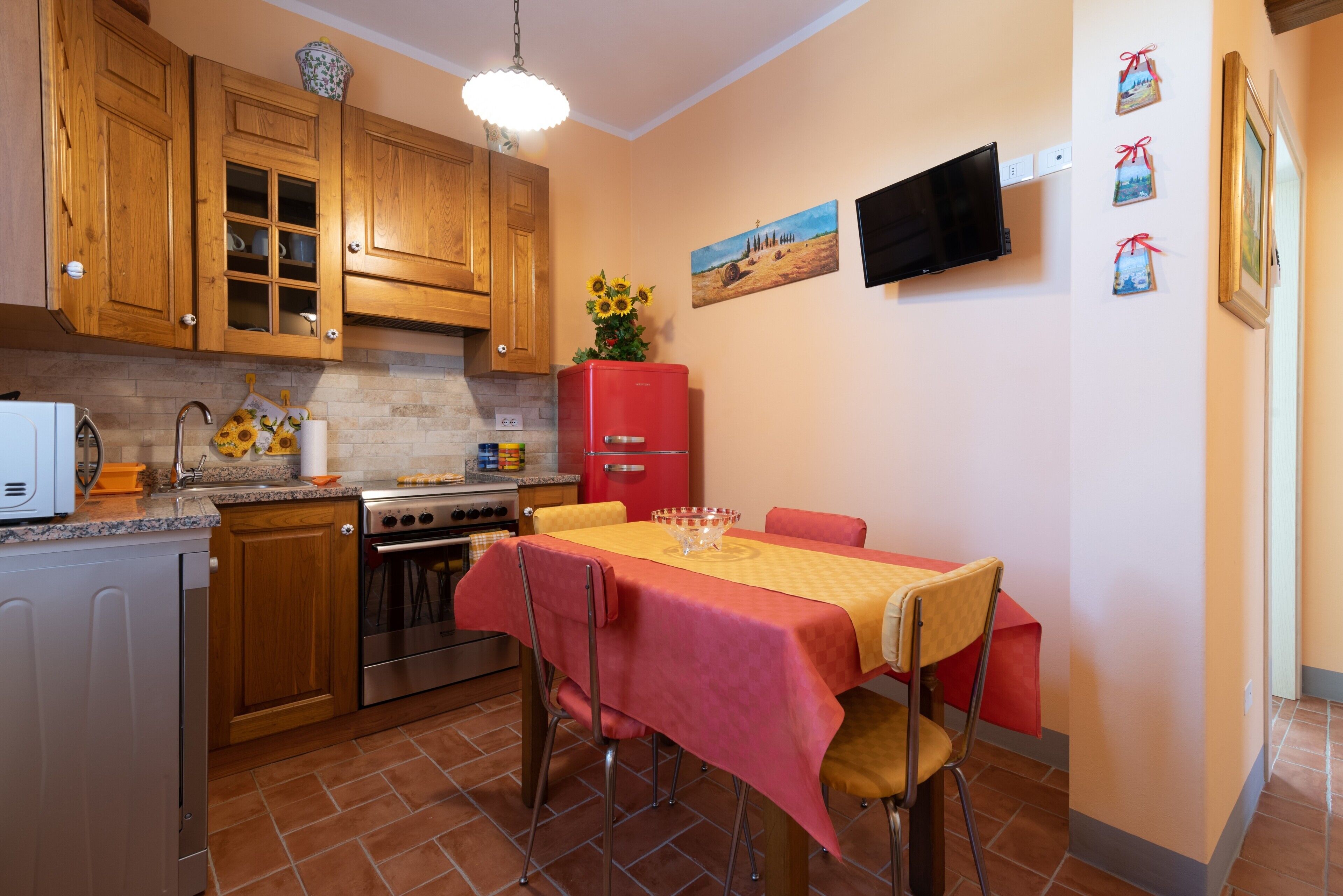 Une cuisine avec des meubles de rangement en bois, un réfrigérateur rouge, une table à manger recouverte d’une nappe jaune et un téléviseur fixé au mur.
