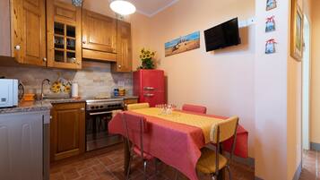 Une cuisine avec des meubles de rangement en bois, un réfrigérateur rouge, une table à manger recouverte d’une nappe jaune et un téléviseur fixé au mur.