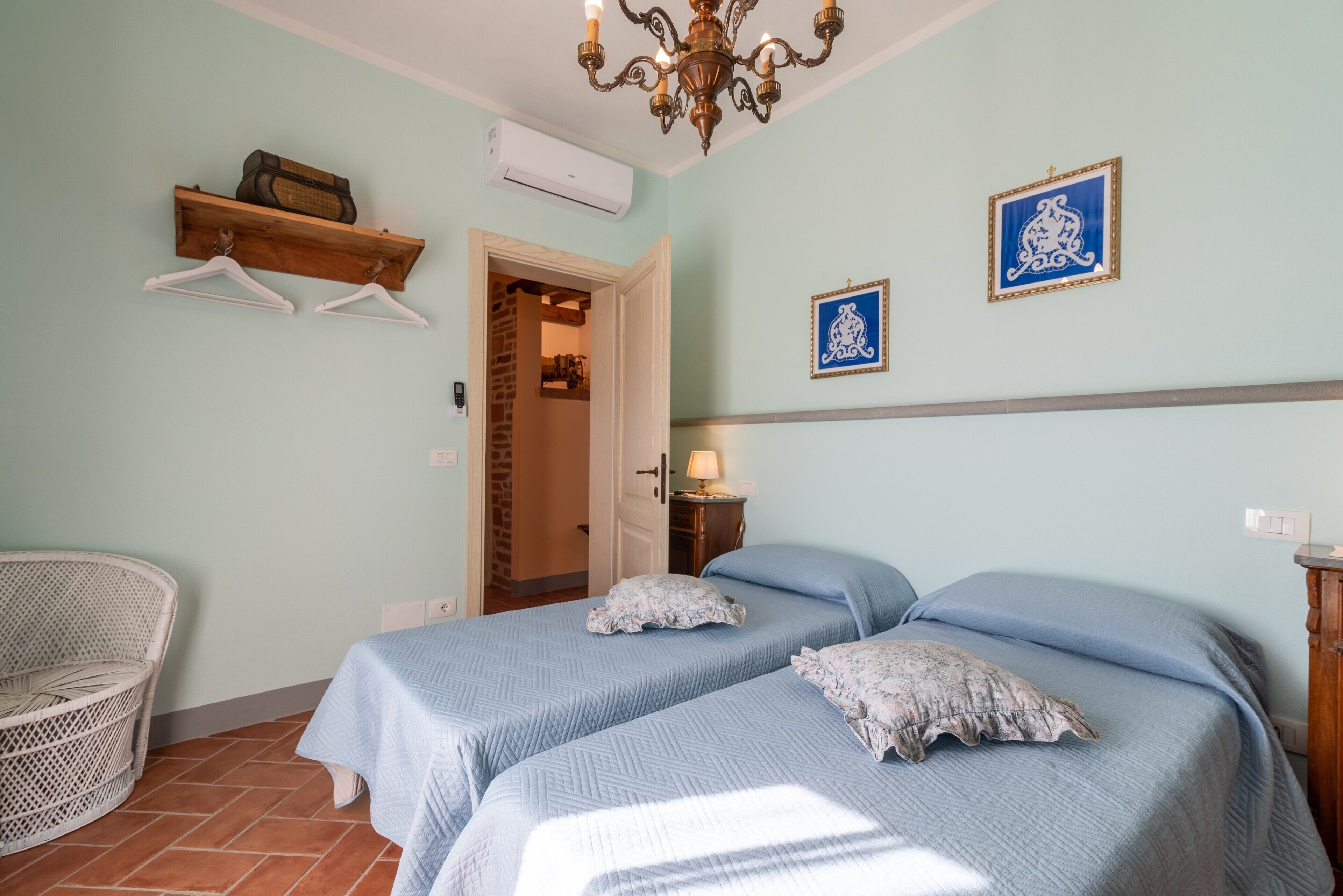 A bedroom with two beds, a wicker chair, a chandelier, and wall-mounted pictures.