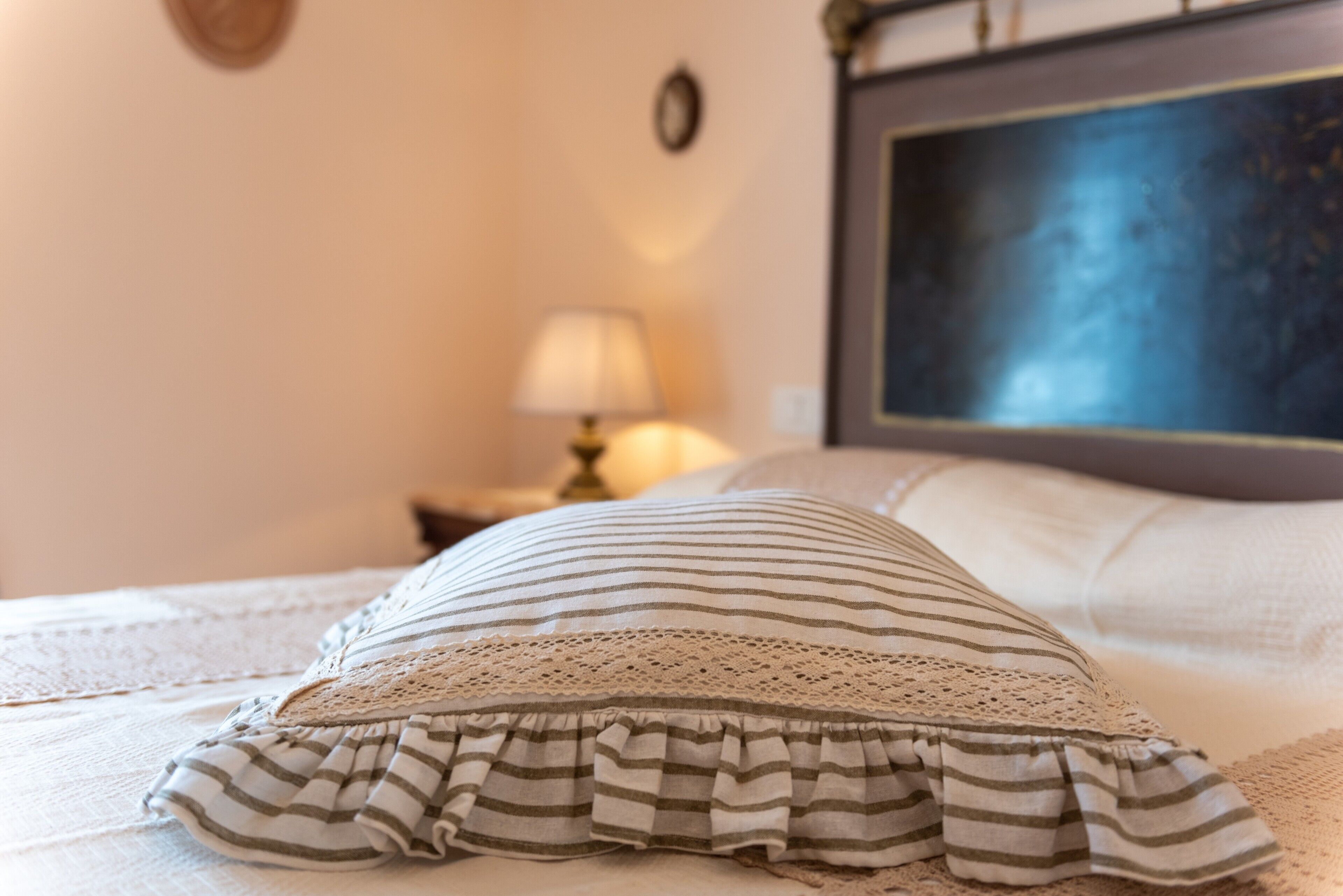 A bed with a striped pillow and a lace-trimmed ruffle, a bedside lamp, and a framed picture on the wall.