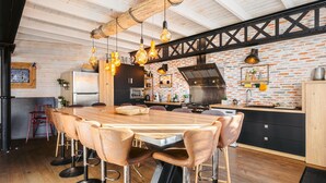 A modern kitchen with a wooden dining table, bar stools, hanging pendant lights, and a brick wall backsplash. - 'Villa la Forge Expérience Bien-Être' with Pool & Wi-Fi (Nuaillé-d'Aunis)