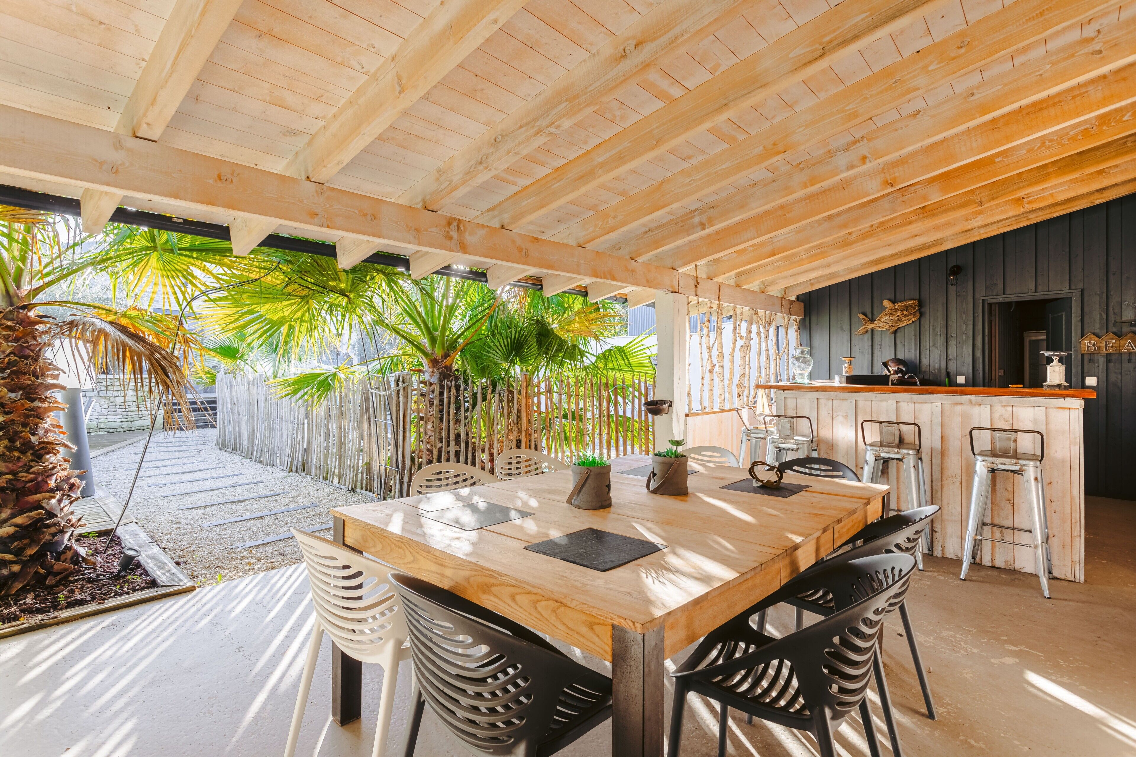 Un espace repas en bois avec une table et des chaises, un coin bar avec des tabourets et des plantes tropicales.