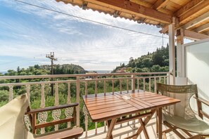 A wooden balcony with a table and chairs, overlooking a lush landscape and a distant view of the sea.