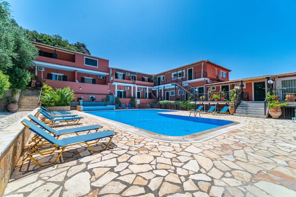 Una piscina con sillas de descanso, un deck con baldosas de piedra y un edificio rojo con balcones.