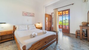 Une chambre avec un lit en bois, des tables de chevet, une commode, un bureau en bois, une chaise et une vue sur l’extérieur par une porte.