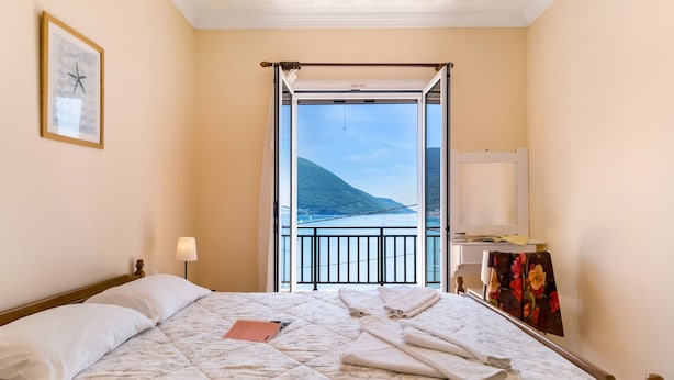 Une chambre d’hôtel comprenant un lit, une lampe de chevet, un tableau encadré, une fenêtre donnant sur une vue de montagne et de l’eau, et un balcon avec une balustrade.