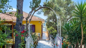 Un chemin mène à un bâtiment jaune, surmonté d’un toit en tuiles, et entouré d’arbres et de plantes.