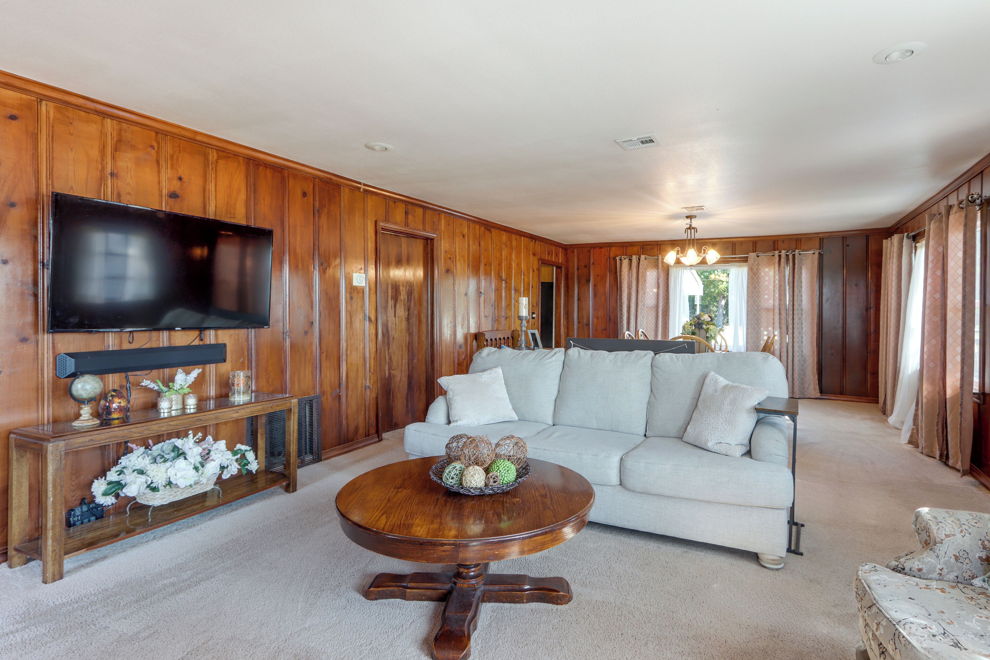 A living room with a wooden paneled wall, a flat-screen TV mounted above a wooden console table, a light-colored sofa, a round wooden coffee table, and a chandelier.