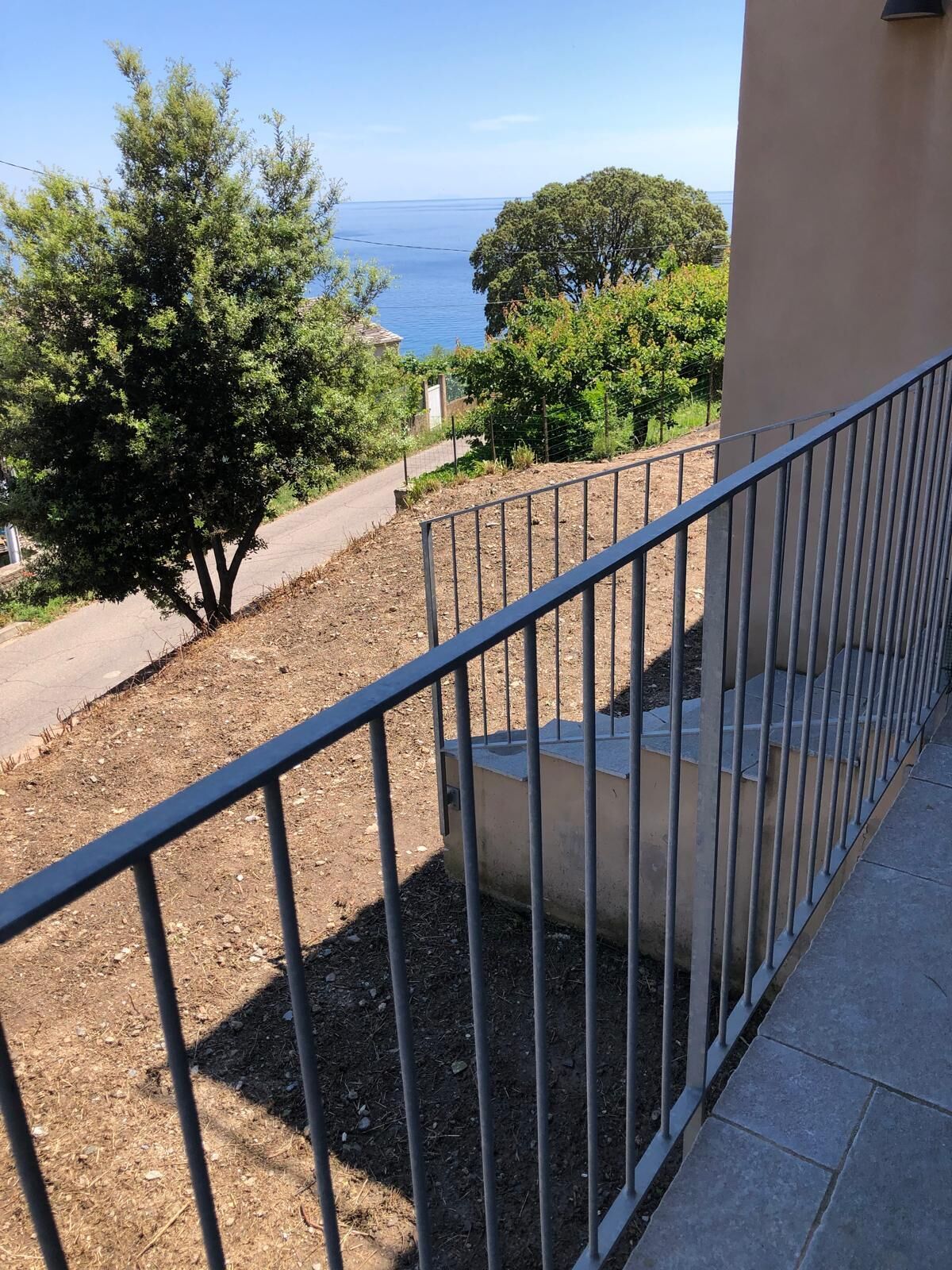A balcony with a metal railing overlooking a dirt path and a tree.