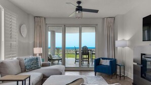 A modern living room with a sofa, ottoman, and a blue armchair. There is a ceiling fan, a lamp, and a fireplace. The room has large glass doors leading to a balcony with a view of the ocean.