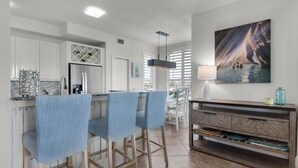 A modern kitchen with white cabinets, a bar area with striped chairs, a wooden dining table, and a large painting on the wall.