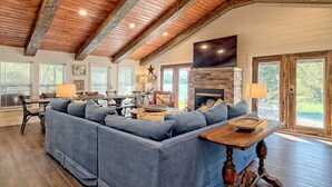 A spacious living room with a large blue sectional sofa, a stone fireplace, wooden beams on the ceiling, and large windows offering a view of the outdoors.