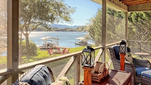 A covered outdoor seating area with wicker furniture, two lanterns, and a view of a lake with dock and chairs.