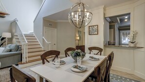 A dining area with a white table set for four, surrounded by wooden chairs. A chandelier hangs above the table, and a staircase is visible in the background.