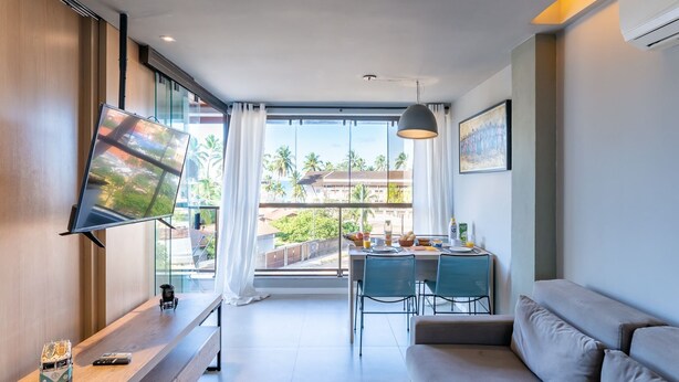 A modern living room with a large TV, a dining area with a table and chairs, and a sofa. - Praia Central Porto de Galinhas (Ipojuca)