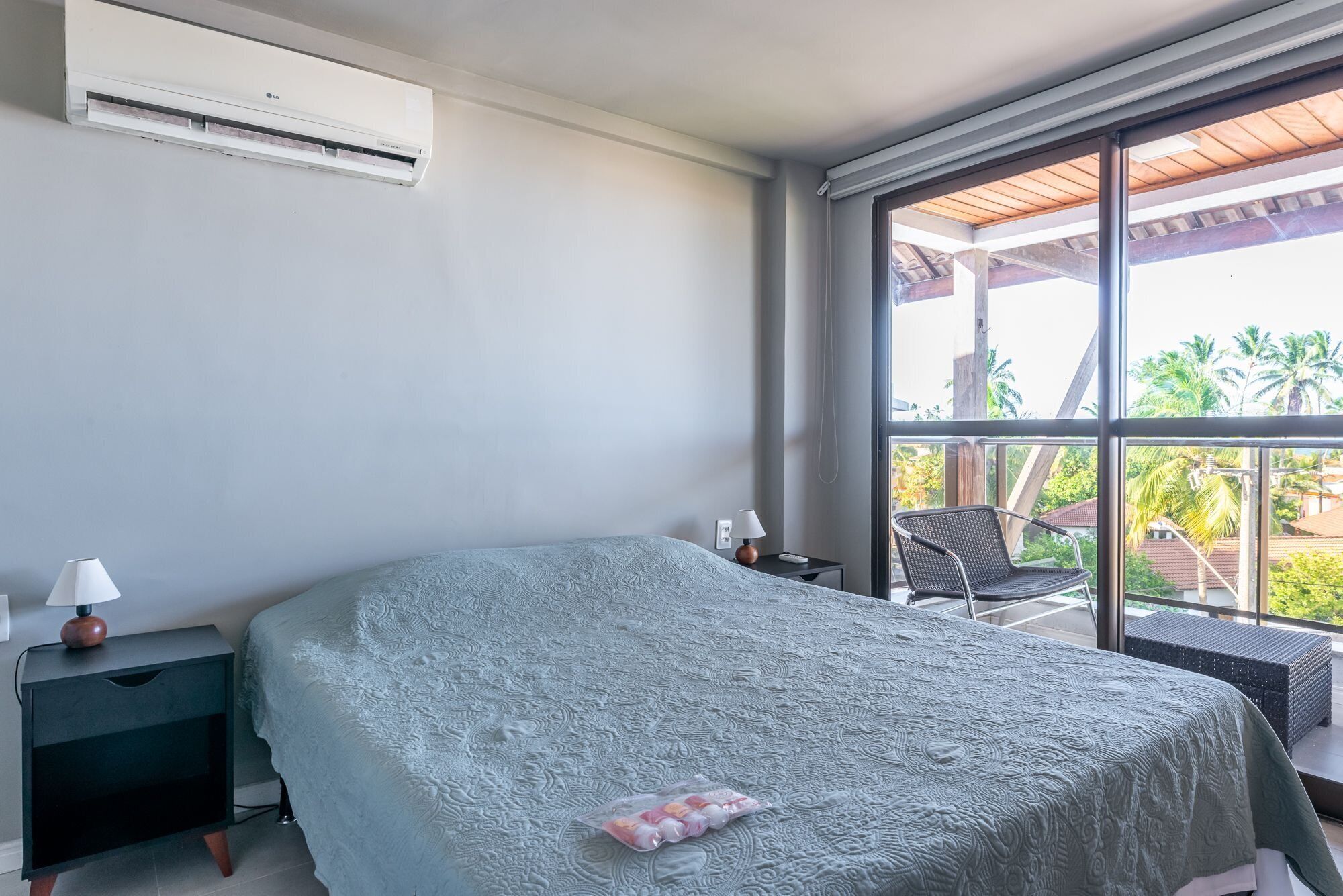 A bedroom with a bed, a nightstand, a lamp, and a window with a view of palm trees.